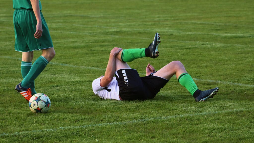 Ein Fußballspieler liegt verletzt auf dem Rasen. Neben ihm steht der Fußballer, der ihn gefoult hat. Symbolbild: Private Haftpflichtversicherung bei maxXse Versicherungsmakler in Nürnberg.
